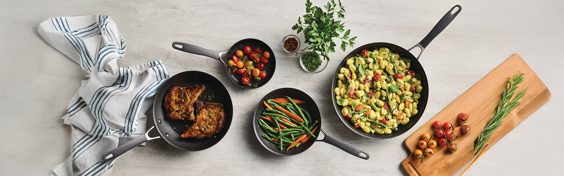 Four pans with cooked food, including meat, green beans, tomatoes, and gnocchi with vegetables; a striped towel, cutting board with tomatoes and chives, and a parsley plant are nearby.