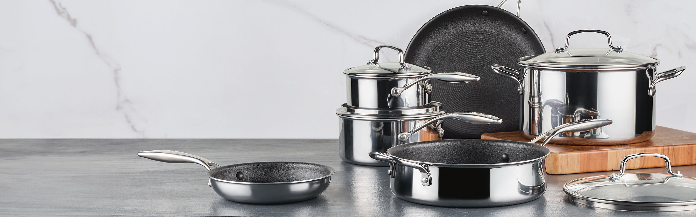 A set of stainless steel cookware, including pots, pans, and lids, is arranged on a kitchen counter with a wooden cutting board.