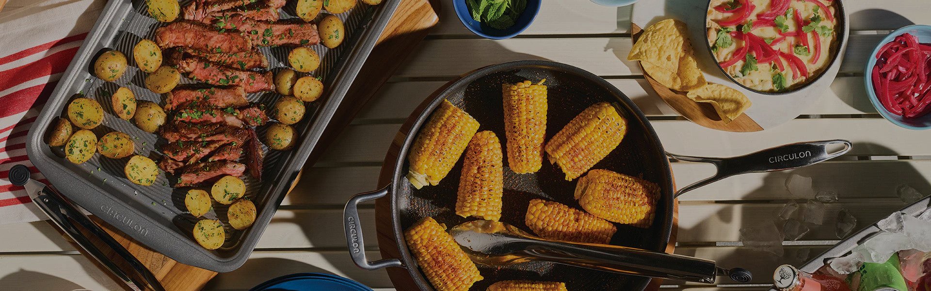 Grilled steak slices with potatoes in one pan and grilled corn in another, surrounded by various side dishes on a picnic table.