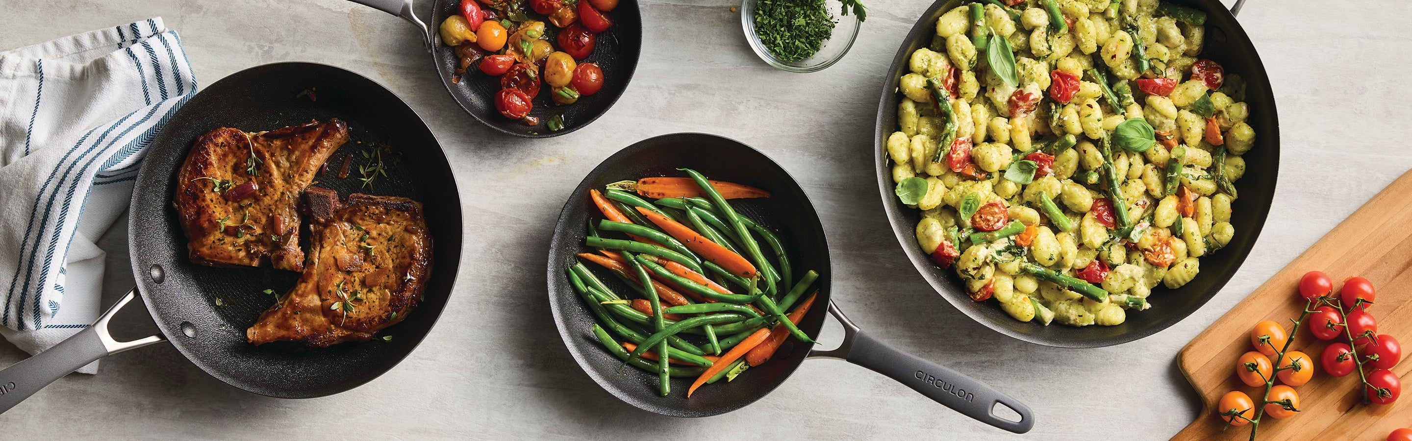 Assorted kitchen cookware and utensils, including pots, pans, a roasting rack, muffin tin, spatulas, measuring cups, and vegetables, arranged on a dark surface.