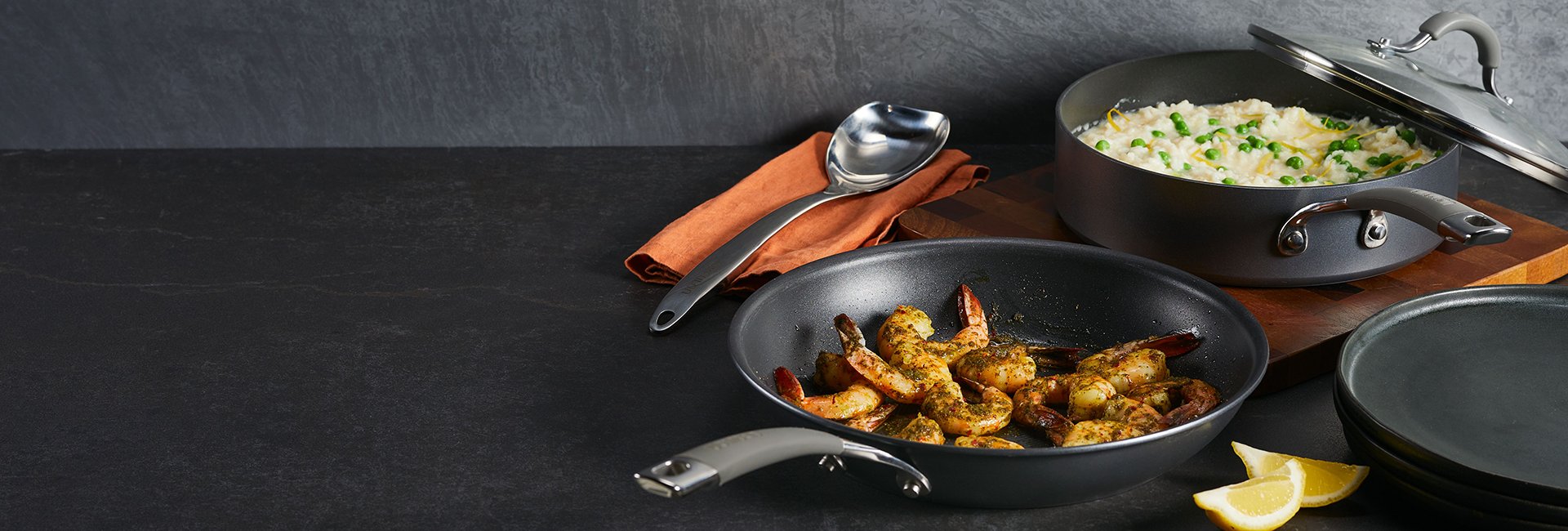 Two nonstick pans on a dark surface: one with seasoned shrimp, the other with rice and peas. A serving spoon, napkin, lemon wedges, and plates are nearby.
