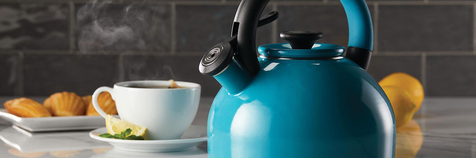 A blue kettle on a countertop next to a steaming cup of tea with a lemon slice, and pastries on a plate.