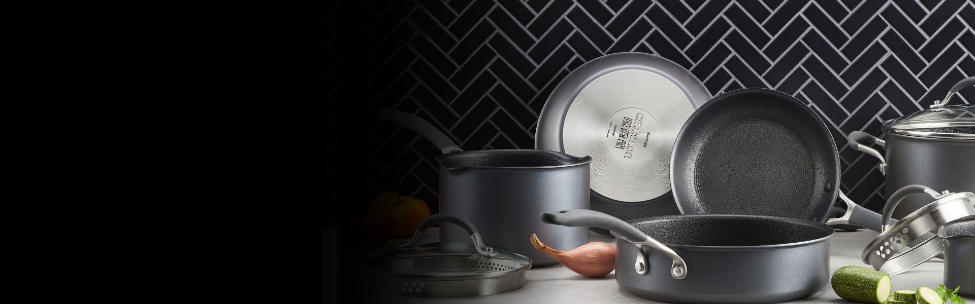 Assorted nonstick pots and pans with lids are displayed on a kitchen counter in front of a black herringbone tile backsplash.
