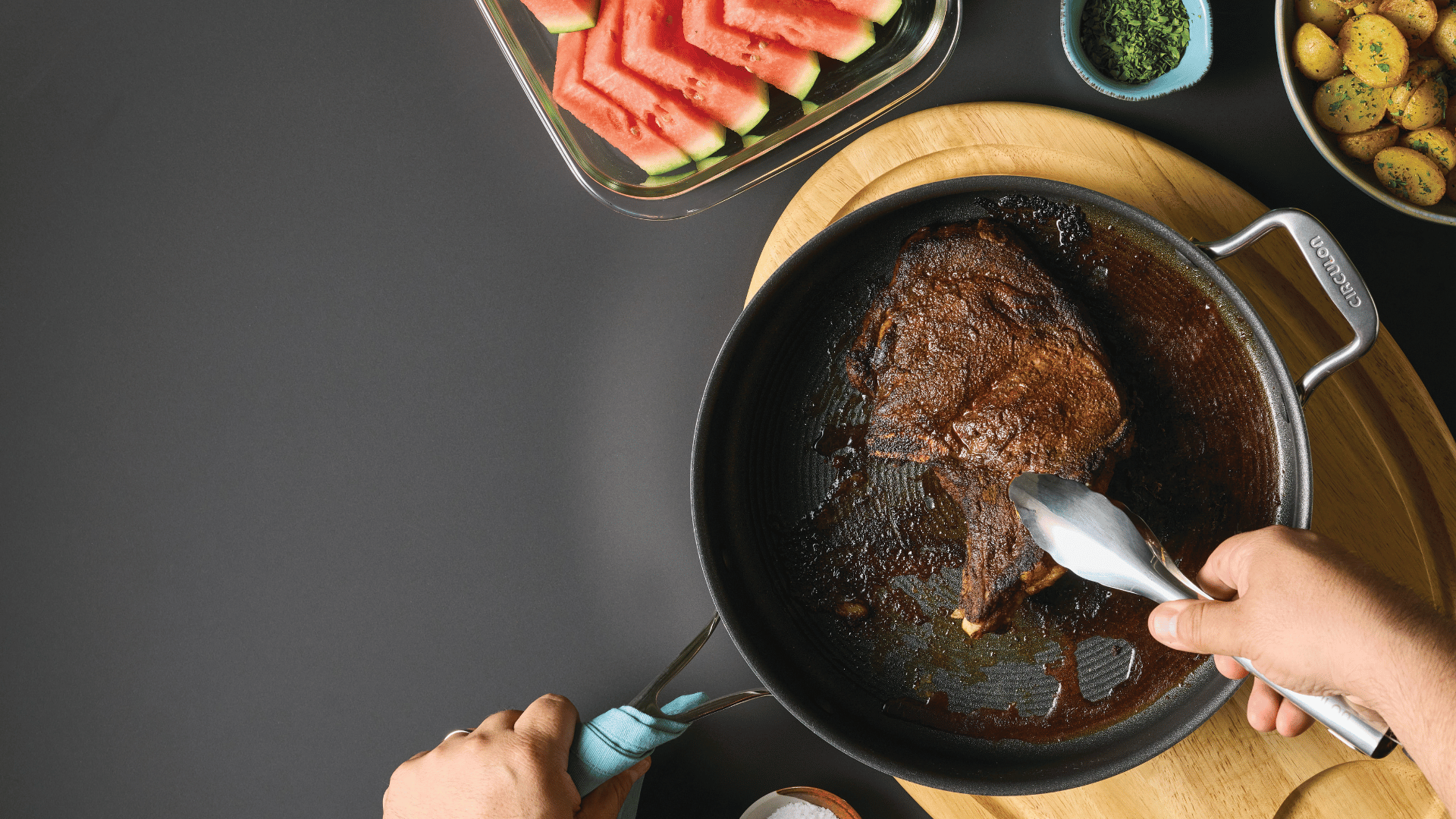 Person holding a pan with a cooked steak using tongs; watermelon slices, small potatoes, and seasoning are on the table.