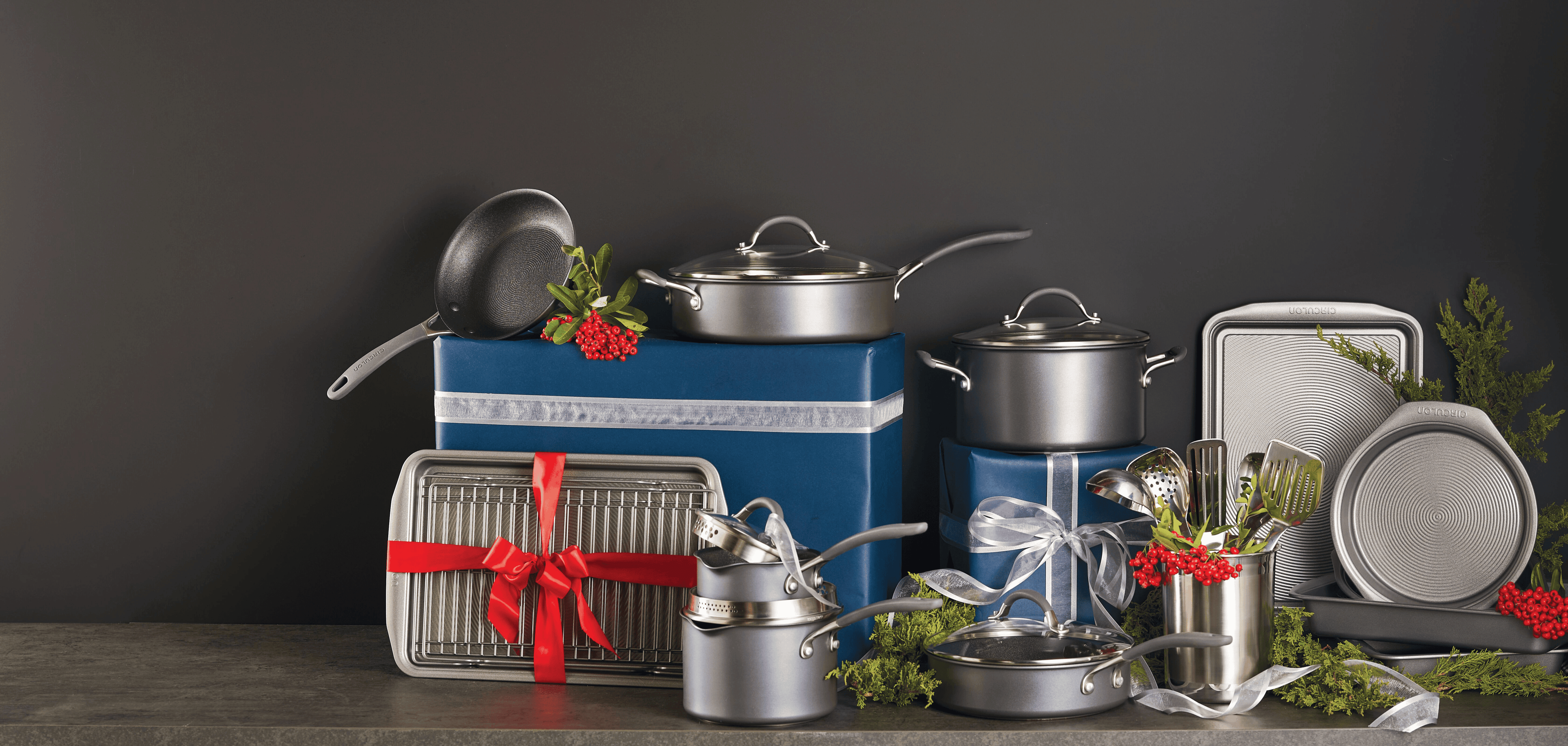 Assorted pots, pans, and kitchen utensils arranged with blue gift boxes, red ribbons, and greenery against a dark background.