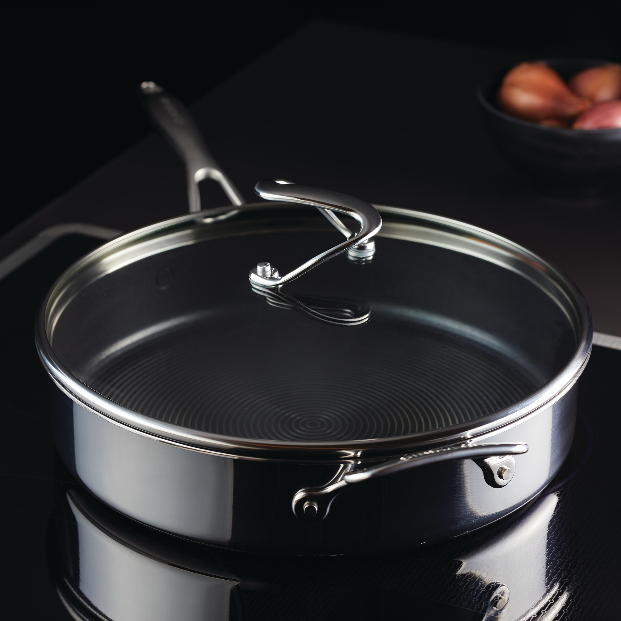 Stainless steel pan with handle and lid on a stove; bowl of shallots in background.