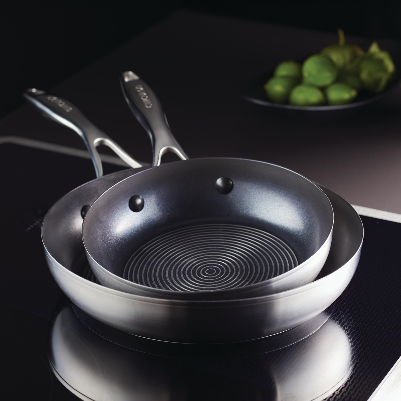 The 8" & 10.25" Hybrid Stainless Steel Open Frying Pans are elegantly stacked on a stovetop with green fruit in the background, creating a harmonious kitchen scene.